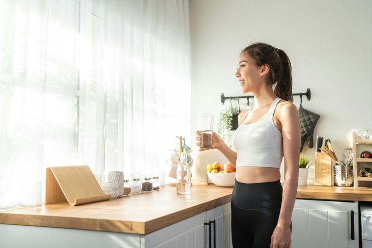 Bright young woman enjoying a glass of water in a modern kitchen with natural light, healthy lifestyle, and clean space.