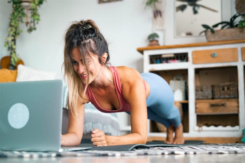 Ejercitamiento en casa, mujer haciendo plancha en su sala con laptop, entrenamiento para pérdida de peso, fitness y bienestar en el hogar.