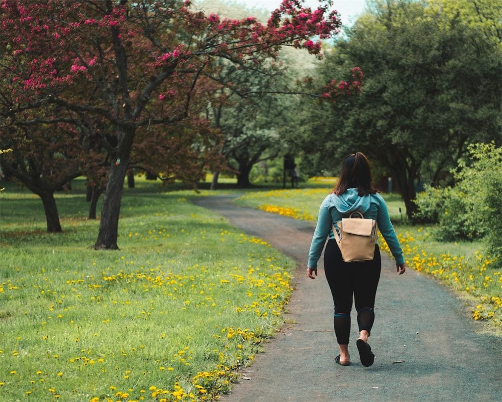 Camino en parque con árboles en flor y mujer caminando con mochila, entorno natural y tranquilo, ideal para actividades al aire libre y relajación, enfoque en naturaleza y paseo.