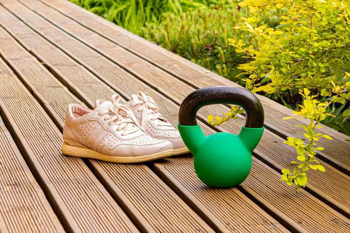 Zapatillas deportivas blancas y kettlebell verde en una terraza de madera rodeada de vegetación.